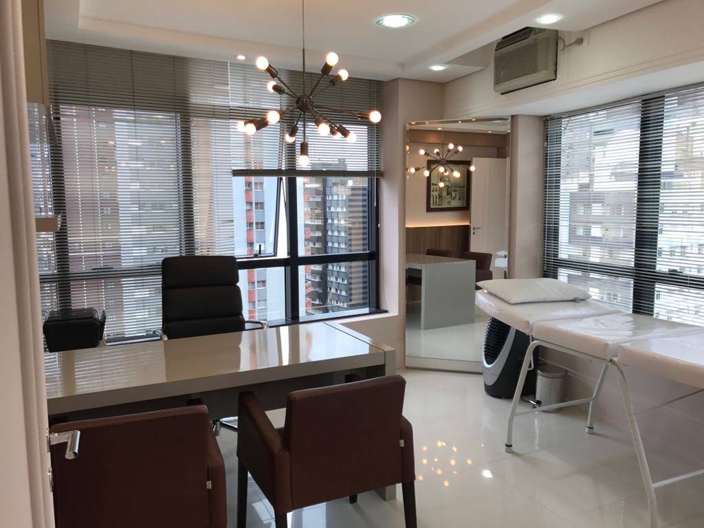 Sala de consulta da Dra. Flávia, com mesa, cadeiras e maca junto às janelas.