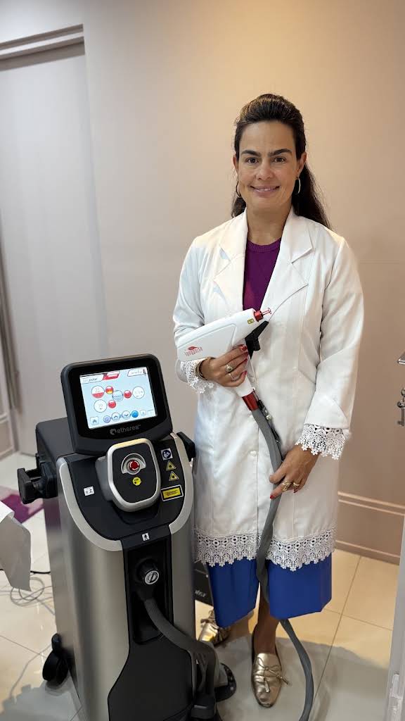 Dra. Flávia ao lado do equipamento laser Etherea, segurando seu aplicador no consultório.